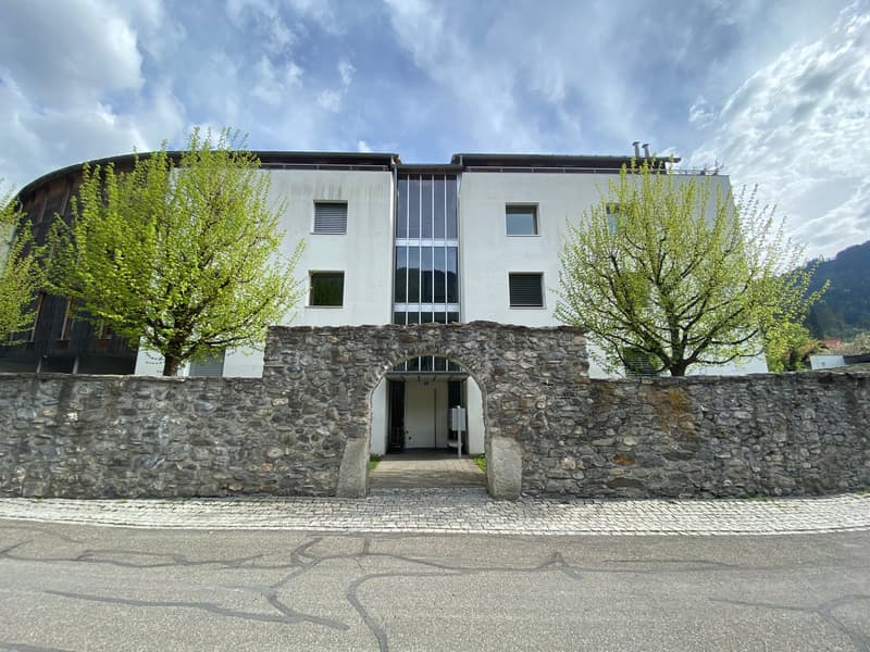 A white building with glass windows and a stone archway at the entrance, two trees are on either side of the archway, a cobblestone pathway leads to the building's entrance, and there is another building in the distance with a sloped roof.