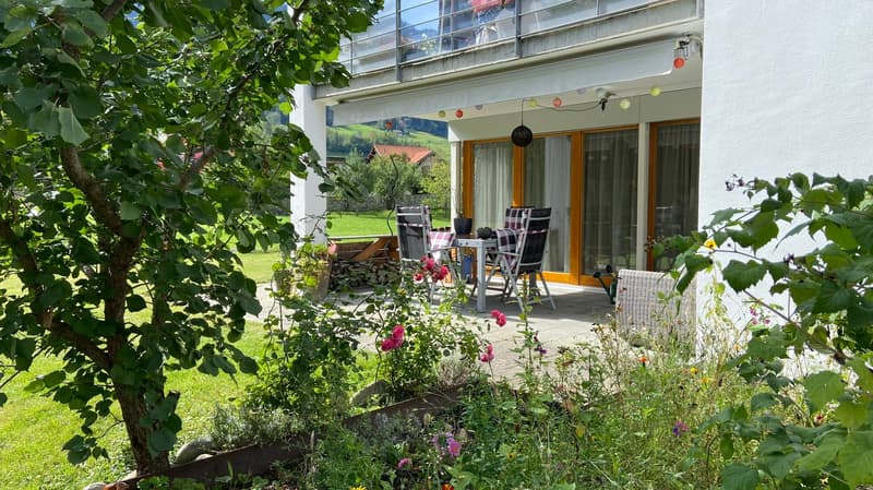 outside area with table and chairs, garden, balcony with railings, green grass and flowers, building with windows and doors, hanging lights