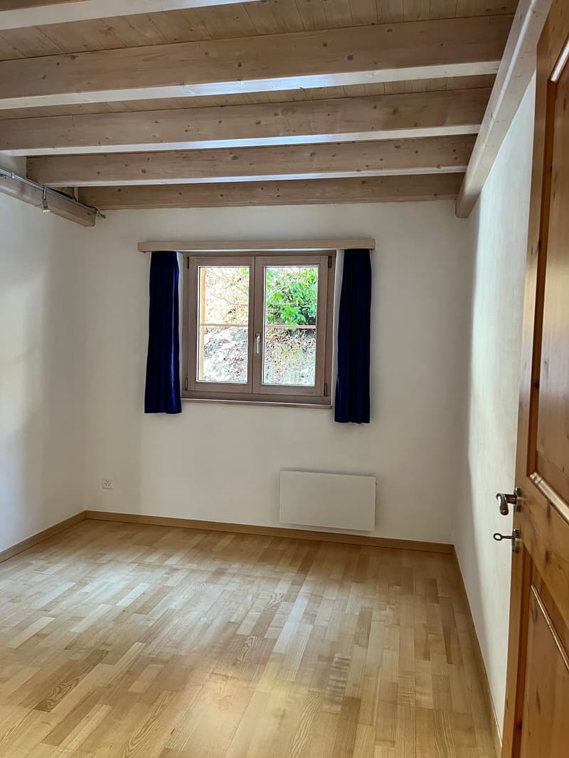 Empty room with wooden floors, white walls, window with blue curtains, wooden ceiling, and a radiator below the window.