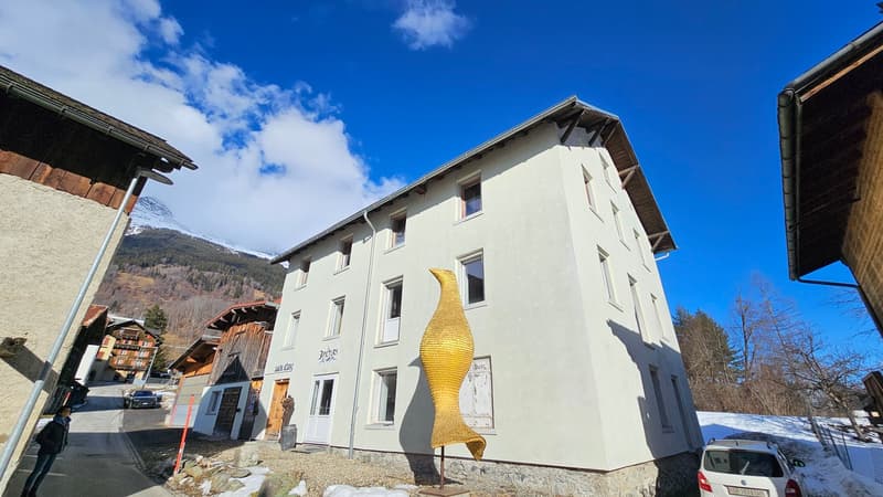White building with 5 windows on the second floor and 5 on the first floor, yellow sculpture on the side of the house, wooden entrance, cars parked in front