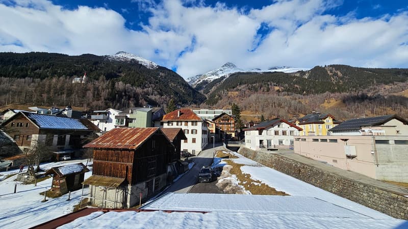 A village with many houses and small buildings, surrounded by snowy mountains and trees. The houses are mostly made of wood and have steep roofs. The roads are covered with snow and there are some vehicles parked. The sky is blue with some clouds.