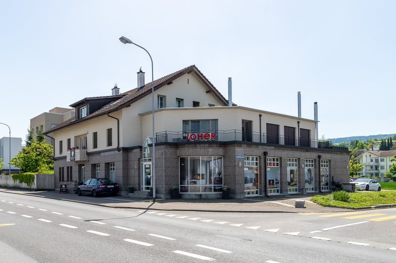 Store building with a name sign reading 'Loher', street-facing facade with glass doors and windows, two cars parked outside, street lighting and greenery in the foreground.