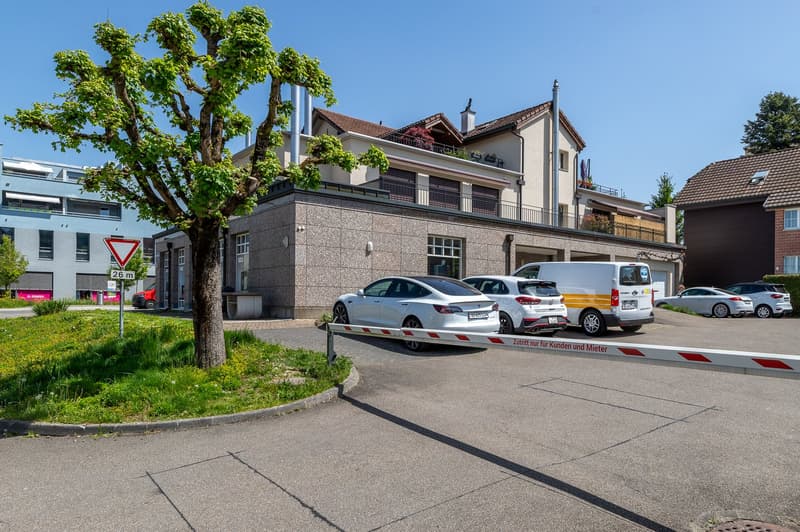 Exterior view of a multistory building with a parking area in front, featuring a tree, grass, parked cars, and a closed gate.