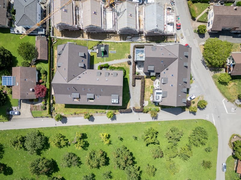 Aerial view of a residential area with several houses, some under construction, surrounded by green grass and trees, with a road and parking area.