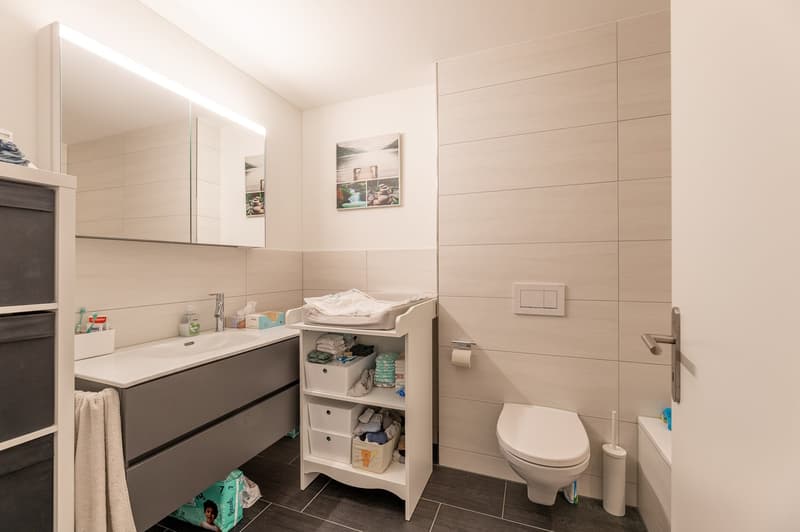 modern bathroom with white tiles, large mirror, white sink, white toilet, white shelves, towel rack, and a small toiletry basket