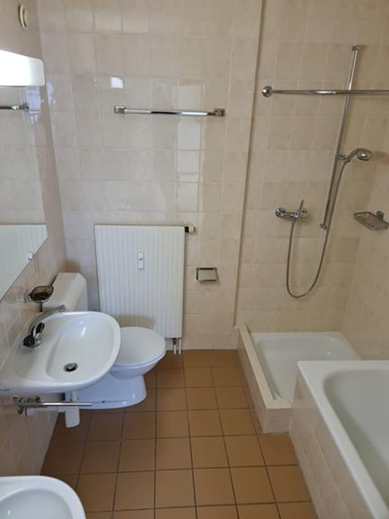 The image shows a bathroom with a sink, toilet, and a bathtub with a shower. The walls are tiled, and there are towel racks and other bathroom fixtures visible.