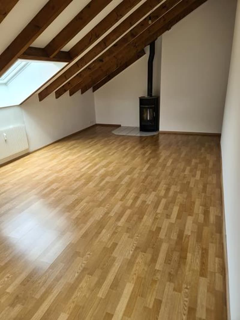 Attic space with exposed wooden beams, hardwood flooring, and a wood-burning stove in the corner.