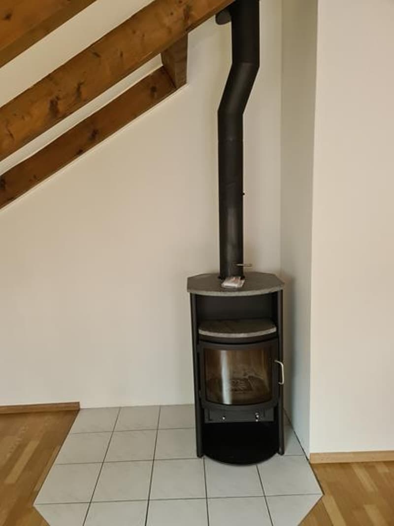 The image shows a wood-burning stove or fireplace in a room with wooden beams on the ceiling and a tiled floor.