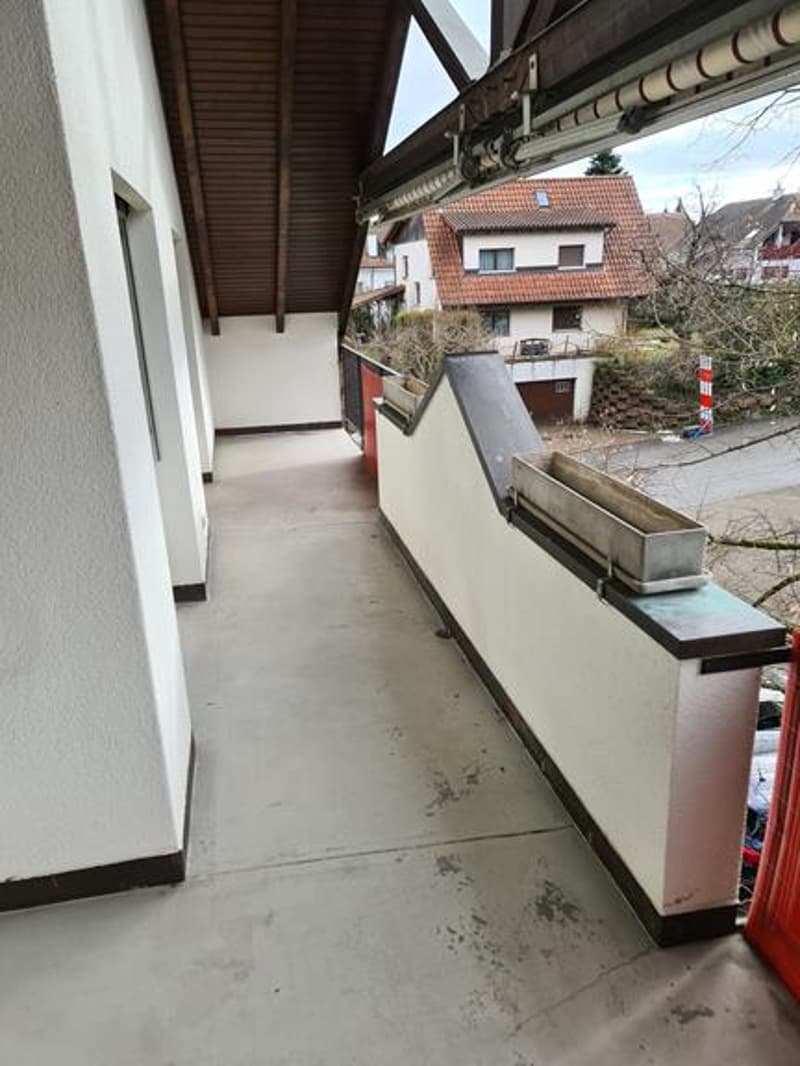 Covered balcony or terrace with concrete floor, overlooking a residential neighborhood with houses and trees in the background.