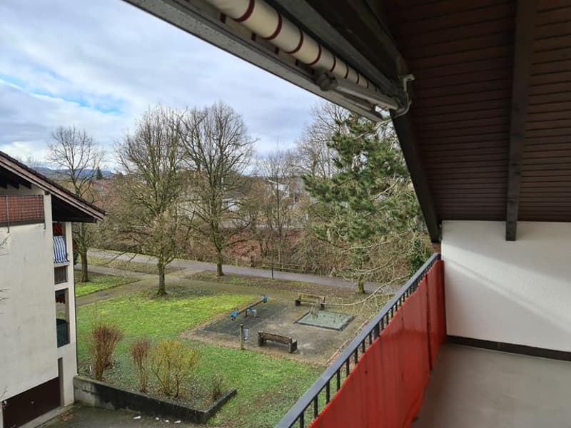 The image shows a balcony or terrace overlooking a grassy area with trees and a bench. The balcony has a wooden roof and railing, and the view outside includes a scenic outdoor space.