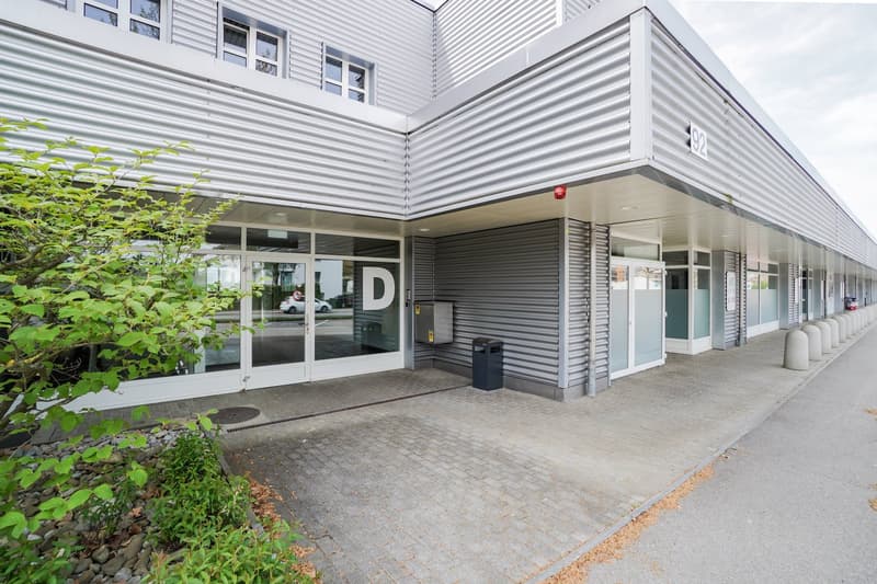 White steel-paneled building with glass doors and windows, bollards, trash cans, brick sidewalk, and parking area.