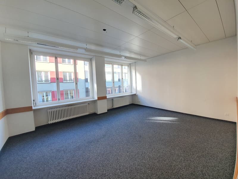 Large open office space with white walls, gray carpeted floor, and multiple windows providing natural light. The room appears to be empty and ready for use.