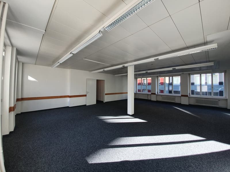 Large open office space with high ceilings, white walls, black carpeted floor, and large windows providing natural light. The room has a clean and modern appearance, with track lighting fixtures installed on the ceiling.