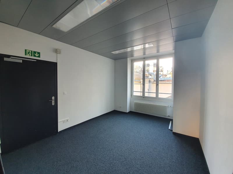 The image shows an empty office space with a gray ceiling, white walls, and a blue carpeted floor. There is a black door on the left side of the room and a large window on the right side, providing natural light. The room appears to be a simple, functiona