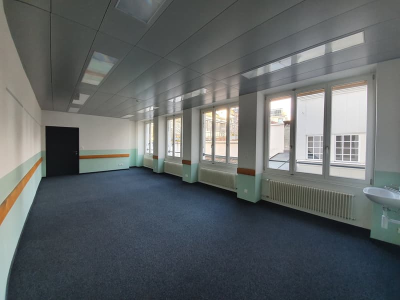 The image shows a large, open office space with a gray ceiling, gray walls, and a blue carpeted floor. The room has several large windows along the walls, providing natural light. There is a black door visible in the corner of the room.