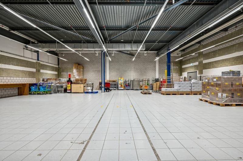 Warehouse with tiled flooring, large open space, several storage items and pallets, metal beams on ceiling, blue support columns, trolleys, trolleys with products, yellow markings on floor.
