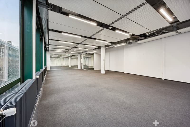 Empty room with large windows, gray floor, and white walls. The ceiling has a grid structure with recessed lights. There are radiators on the left side, and a white ceiling above.
