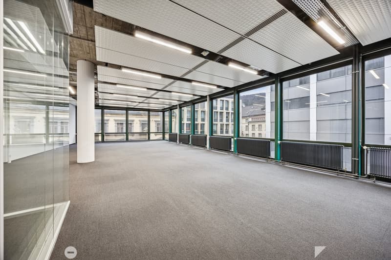 Empty office with large glass windows, carpeted floor, modern ceiling and large pillars.