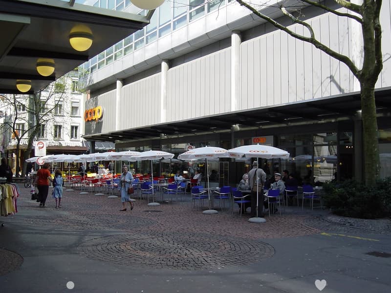An outdoor seating area with tables and chairs under white umbrellas at a restaurant, surrounded by plants and trees with a brick floor, beside a large building with signage and an open entrance