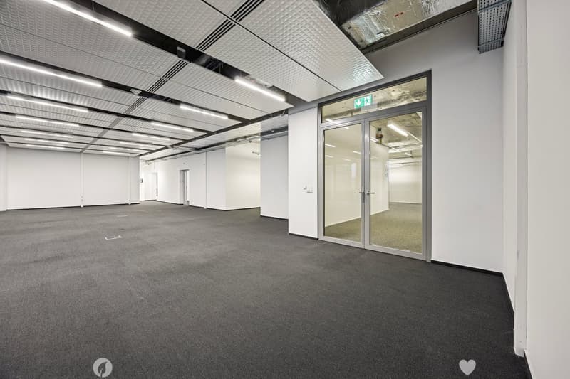 spacious room with gray flooring, white walls, glass doors and windows, ceiling lights, exit sign