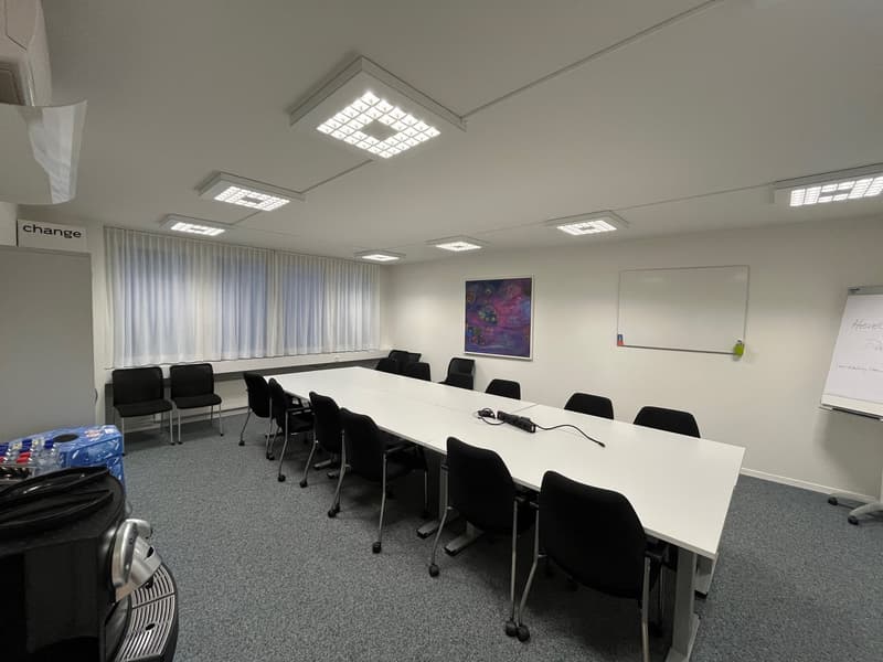 Empty room, large conference table, black chairs, whiteboard, picture on wall, ceiling lights, grey carpet, side tables with trash bins