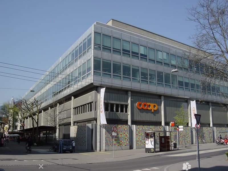 A large office building with glass walls, featuring a large white sign with the word 'COOP', street level shops, a small bus stop, and trees along the street.