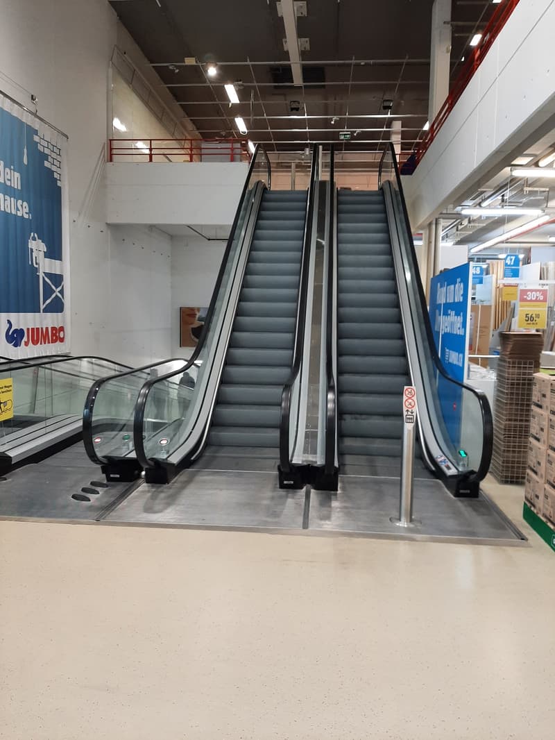 The image shows the interior of a retail store, with two escalators in the center of the frame. The escalators have gray steps and are surrounded by a white and blue tiled floor. The ceiling is high and has various lighting fixtures installed. There are s