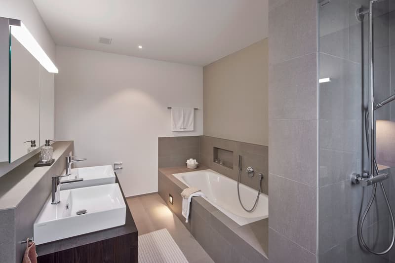 Modern bathroom with white sink, bathtub, and glass shower enclosure. The walls are beige and gray tiles, and there is a towel rack and other bathroom accessories visible.