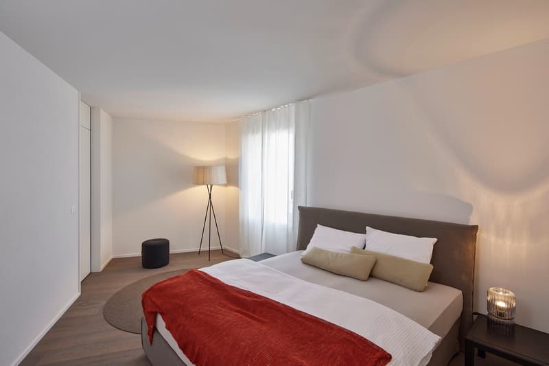 Spacious bedroom with a large bed, white bedding, and a red throw blanket. The room has a floor lamp, a side table, and a curtained window, suggesting a cozy and relaxing atmosphere.