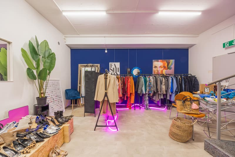 Store with racks of clothes, hanging jacket, shoes and bags, neon lights, plant, table with clothes.