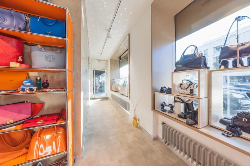 A hall with lots of accessories, designer bags, and shoes. It has wooden floors and is illuminated with spot lights. One wall has a shelf with accessories and bags, and the other has a glass window with designer shoes and bags on display.