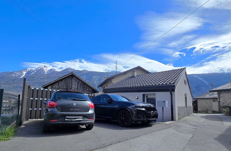 Two cars parked in front of a house with a garage, wooden fence, and mountains in the background