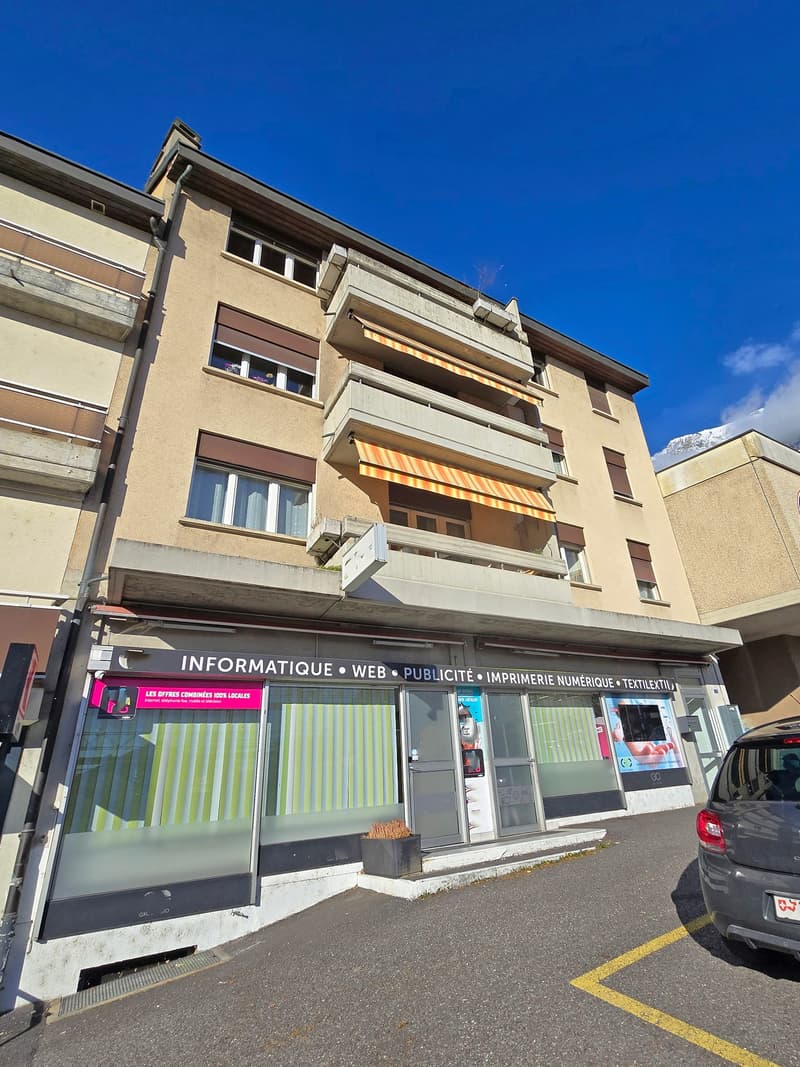 A multi-story building with a commercial space on the ground floor, featuring a sign that reads 'INFORMATIQUE • WEB • PUBLICITE • IMPRIMERIE NUMÉRIQUE • TEXTILEXTIN'. The building has multiple windows and balconies, and there is a car parked in front.