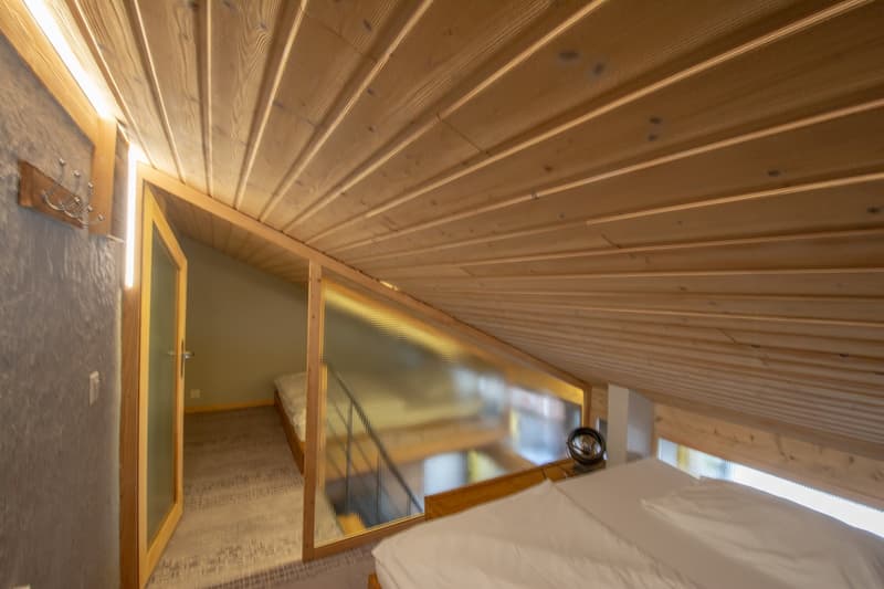 Small bedroom with a bed, slanted wooden ceiling, transparent wall to another room, single bed.