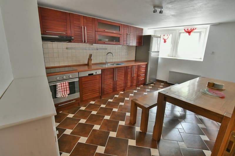 Wooden cabinets, tiled flooring, stainless steel appliances, refrigerator, sink with faucet, small dining table and bench, small window with red curtains, small countertop, towel hanging on the oven handle