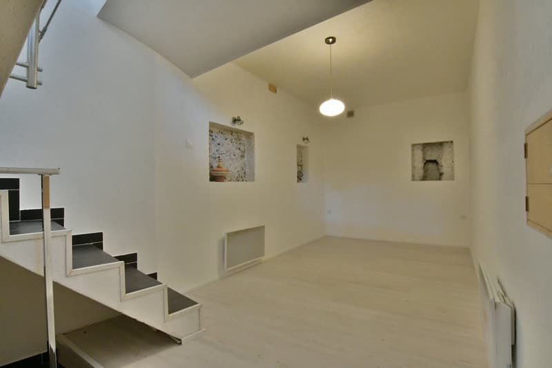 White walls, white floor, pendant lighting, built-in shelf with decorative item, heater on the wall, small window on the right, staircase on the left.