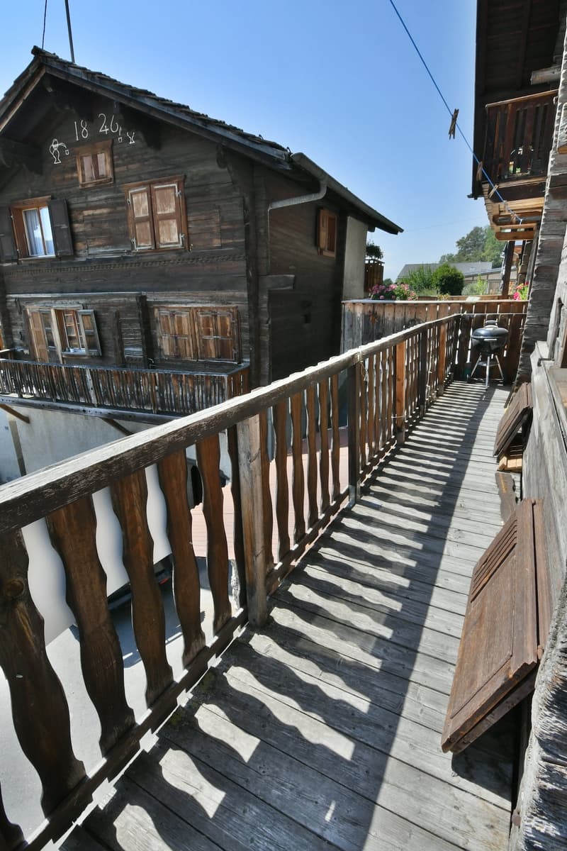 wooden balcony, white car on street, small grill, balcony of building on left, fence on right, shadows on the ground