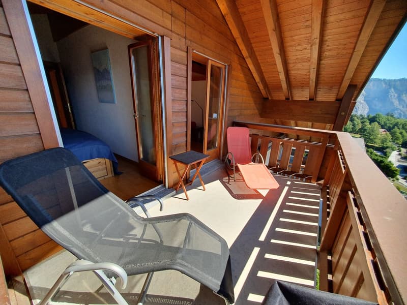 Outdoor wooden balcony with reclining chairs, small table, railings, wooden floor, and view of greenery and mountains.