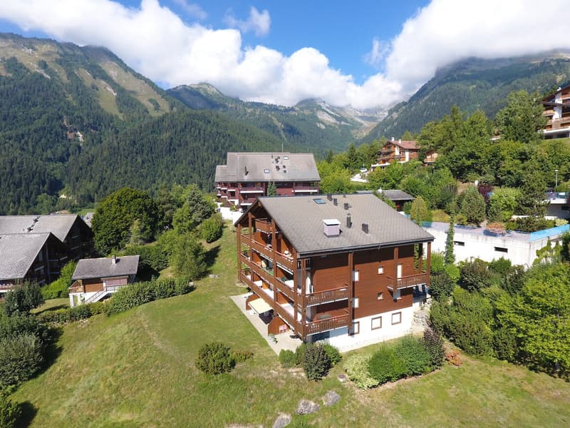 A building with balconies and terraces surrounded by trees and grass, mountains in the background
