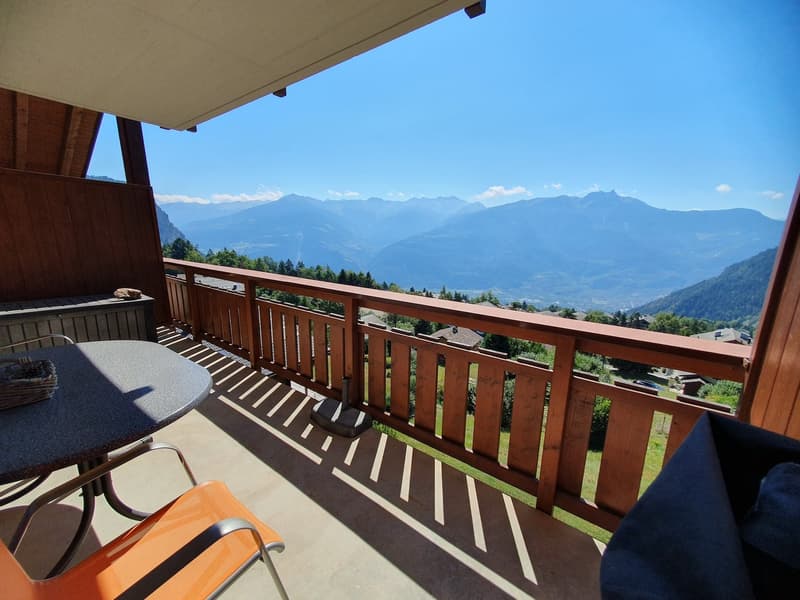 Terrace with wooden railing, round table, chairs, clear sky, mountains in the distance, wooden flooring