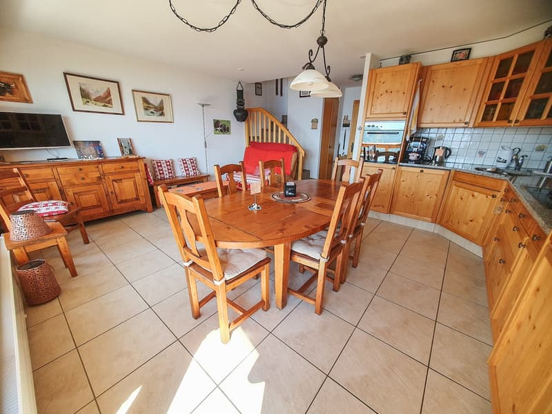 room features a wooden dining table and chairs, tiled floor, wooden cabinets, kitchen area with an oven, picture frames, staircase, television on a stand, and a lamp