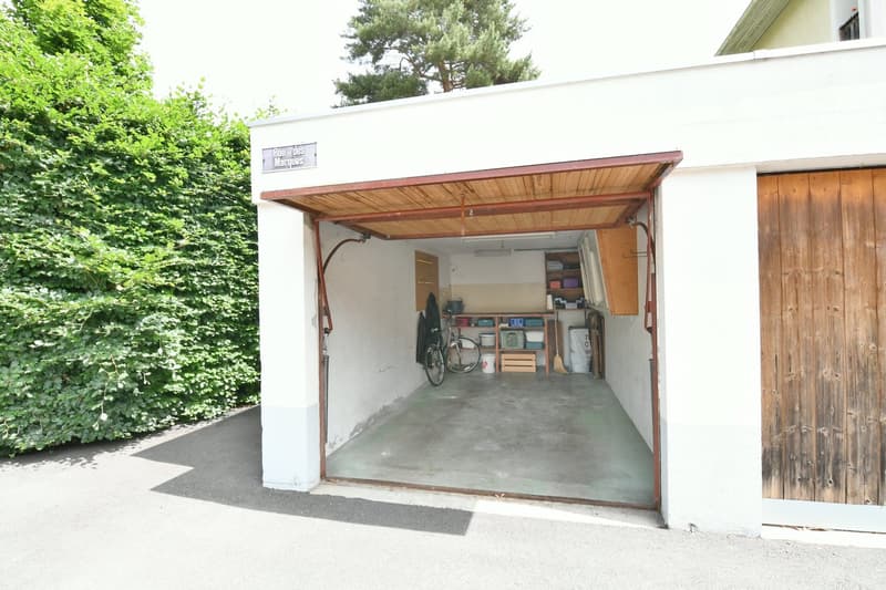 A detached garage with a wooden roof and concrete floor. The garage is filled with various storage items and a bicycle.