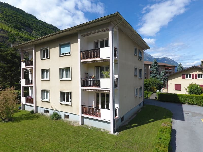 3-story apartment building with balconies, surrounded by greenery and mountains in the background