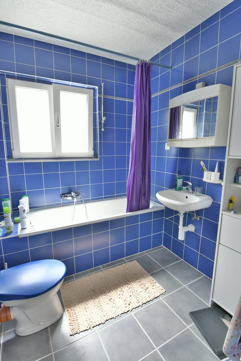 The image shows a bathroom with blue tiled walls, a white sink, a bathtub, and a purple shower curtain. The floor is tiled in gray, and there is a small rug on the floor. The bathroom also has a window and some shelving units for storage.