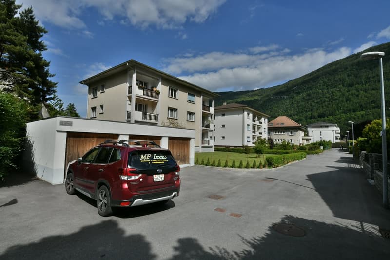 A multi-story apartment building with balconies, surrounded by trees and mountains. A red SUV is parked in the foreground.