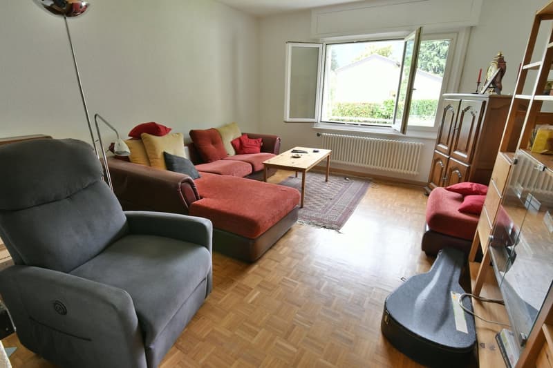 Spacious living room with large windows, hardwood floors, gray recliner sofa, red and yellow accent pillows, wooden coffee table, and shelving unit with decor items