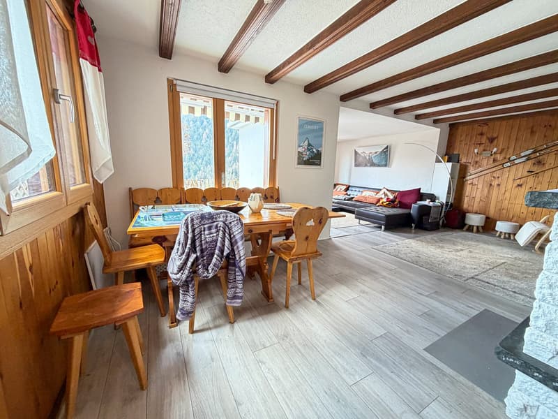 Dining room with wooden table and chairs, wooden beams, white walls, wooden flooring, large window with view, living room with couch and fireplace