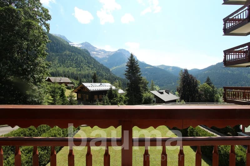 Scenic mountain landscape with forested hills, snow-capped peaks, and traditional wooden chalets or cabins with balconies overlooking the valley