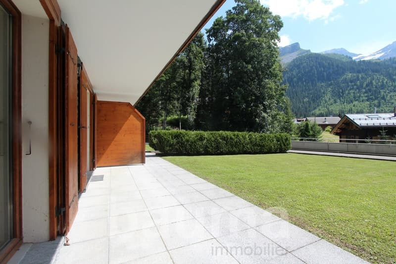 The image shows the exterior of a building with a large patio or balcony area. The patio has a tiled floor and is surrounded by a well-maintained lawn and hedges. The building has a wooden exterior with large windows that offer a scenic view of the mounta