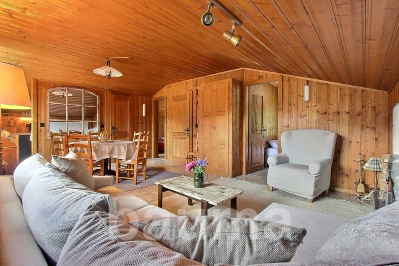 Cozy wooden cabin interior with living room, dining area, and kitchen. The room features wooden walls and ceiling, a fireplace, comfortable sofas and armchairs, a wooden dining table, and various decor elements.
